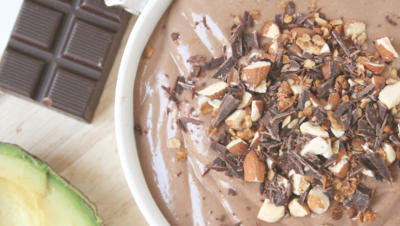 Image of chocolate smoothie bowl with topping of almonds and chocolate, with chocolate and halved avocado on table next to bowl